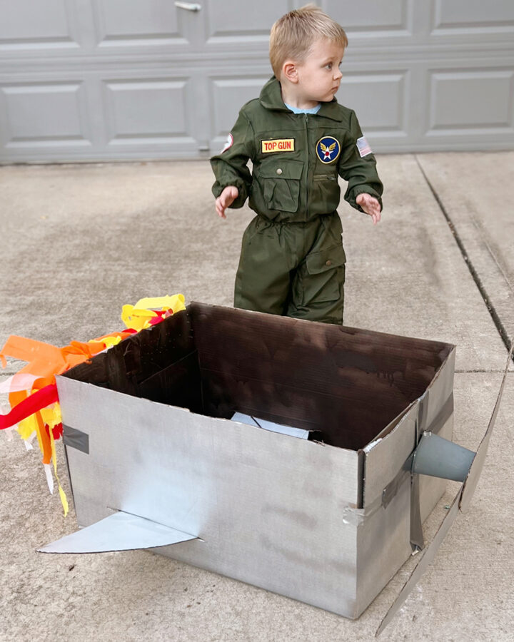 toddler in pilot's costume standing by cardboard box plane.
