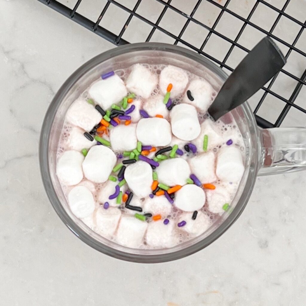 hot chocolate in a glass with marshmallows and purple, green and orange sprinkles. 