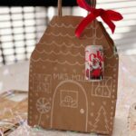 Brown paper bag decorated to look like a gingerbread house with a red ribbon and ornament tied to it.