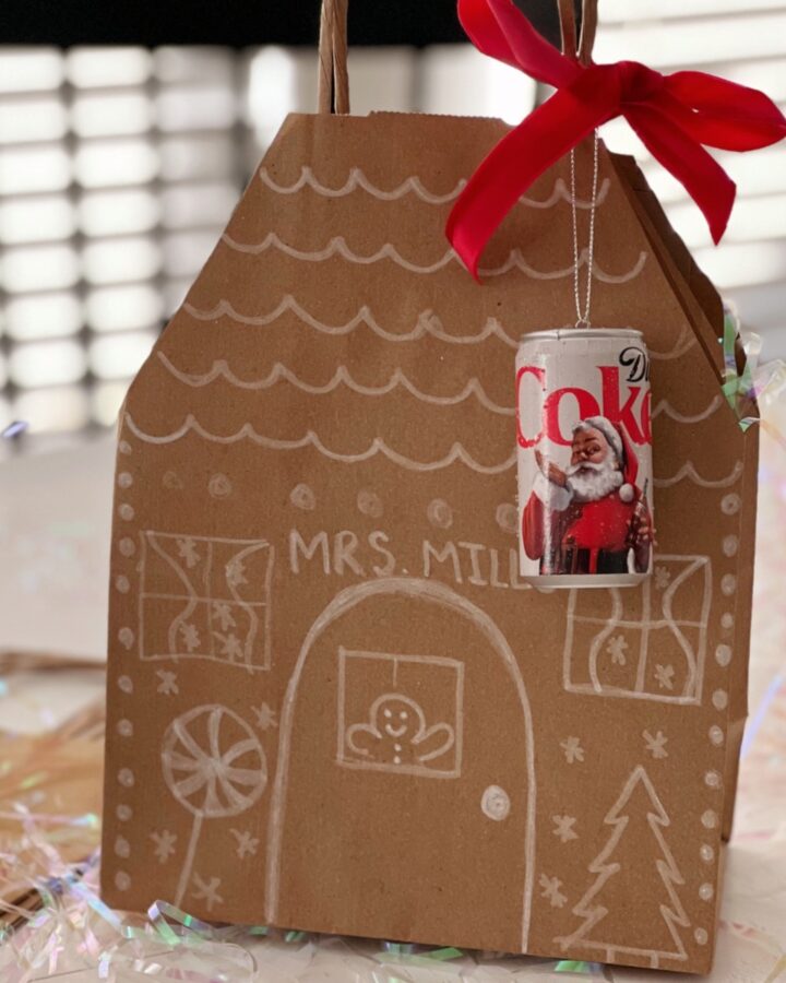 Brown paper bag decorated to look like a gingerbread house with a red ribbon and ornament tied to it.