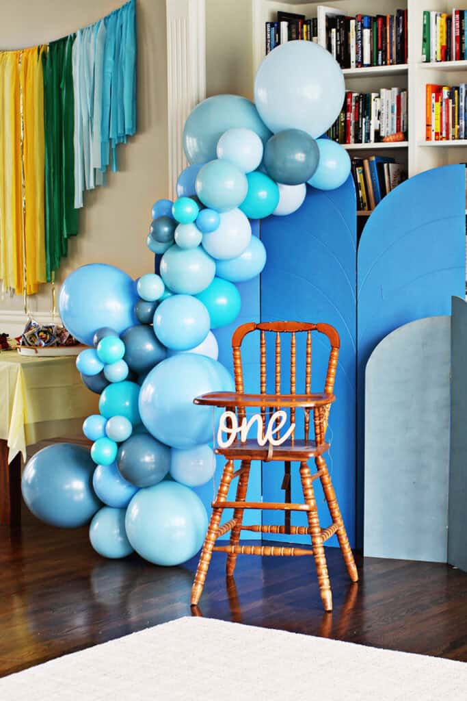 Blue balloon arch around a blue arch backdrop with a wooden high chair in front.