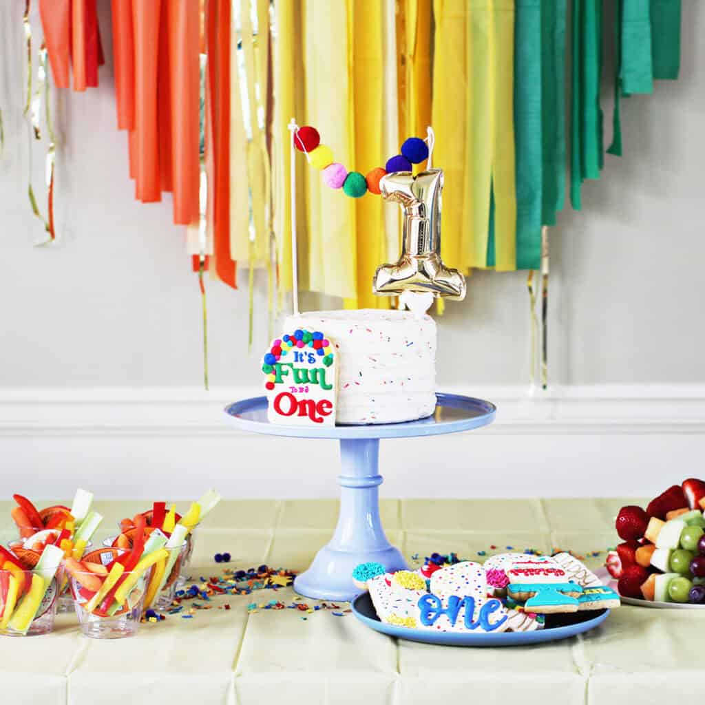 cake on a blue cake stand with a number 1 cake topper. The cake is on a table in front of rainbow streamers. 