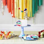 cake on a blue cake stand with a number 1 cake topper. The cake is on a table in front of rainbow streamers.
