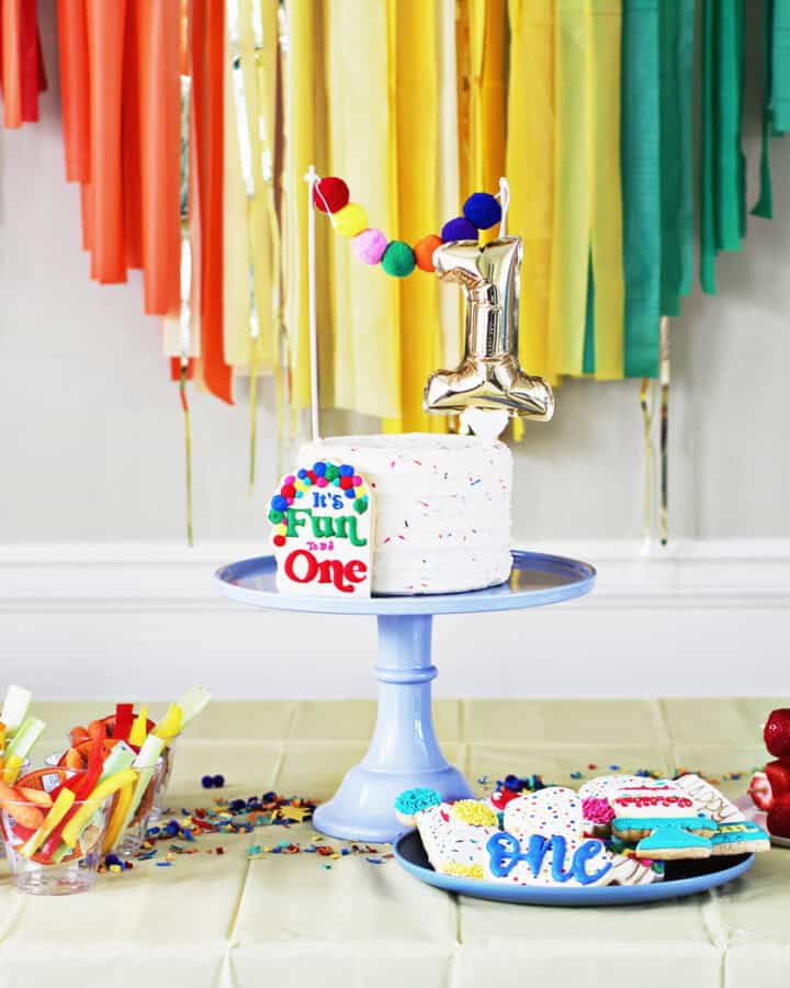 cake on a blue cake stand with a number 1 cake topper. The cake is on a table in front of rainbow streamers.
