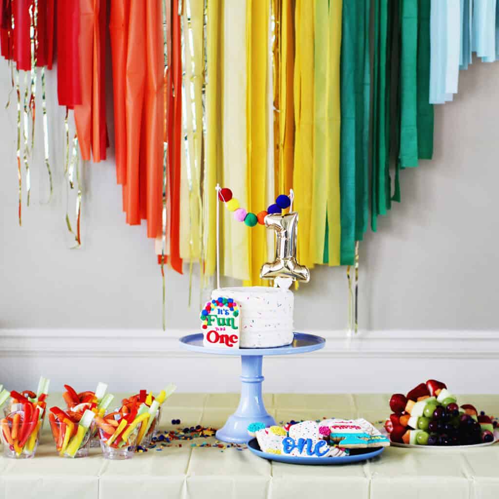 cake on a blue cake stand with a number 1 cake topper. The cake is on a table in front of rainbow streamers.