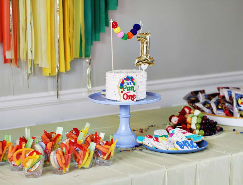 cake on a blue cake stand with a number 1 cake topper. The cake is on a table in front of rainbow streamers.