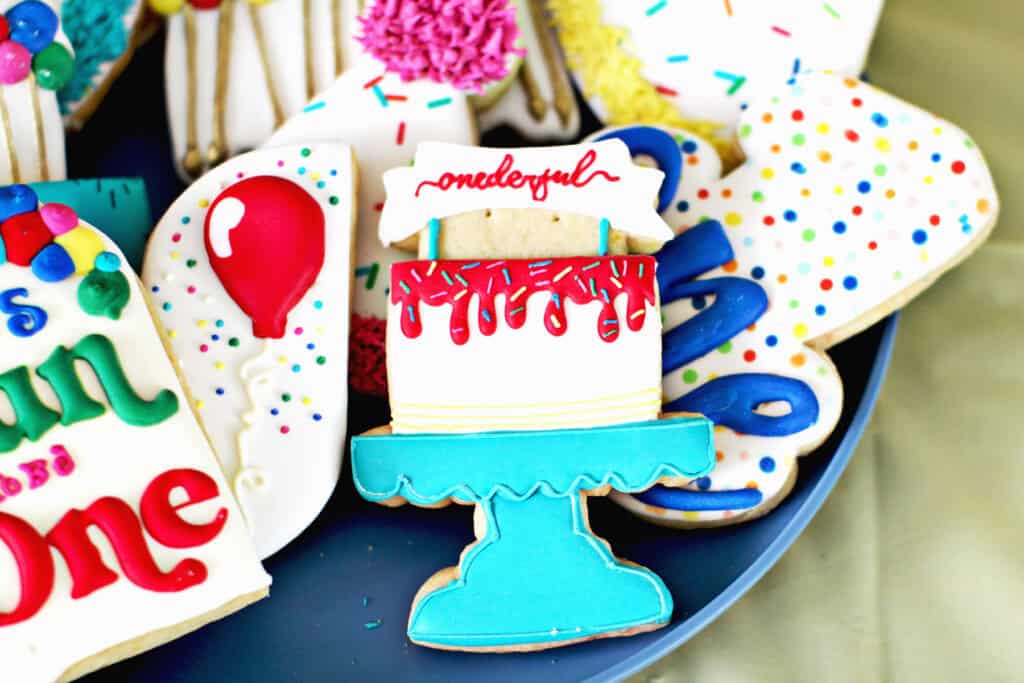Cake on a cake stand sugar cookie on a plate with other birthday themed sugar cookies. 