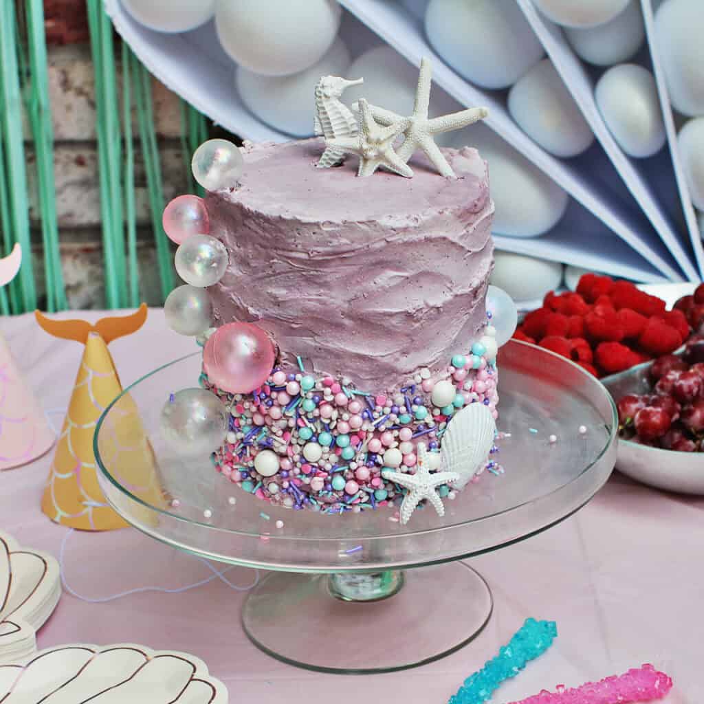 Mermaid-themed birthday cake on clear cake stand.